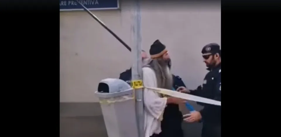 Un bărbat cu barbă lungă și căciulă poartă o haină albă lângă un stâlp metalic, în prezența unui oficial în uniformă întunecată.
