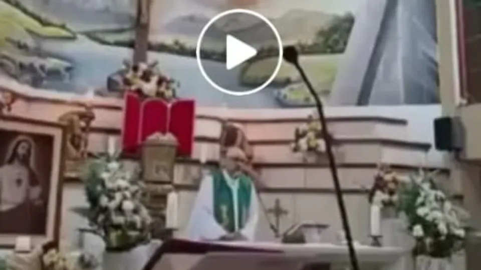 O imagine dintr-un video arată un preot în vestimentații liturgice, în picioare într-un altar decorat cu flori, crucifix și o pictură a lui Iisus.