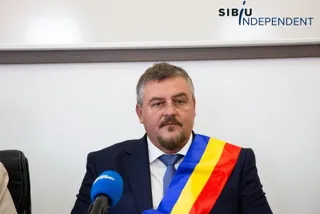 Un bărbat cu barbă și mustață poartă un costum închis, cravată albastră și o bandă tricoloră pe umăr, stând în fața unui microfon.