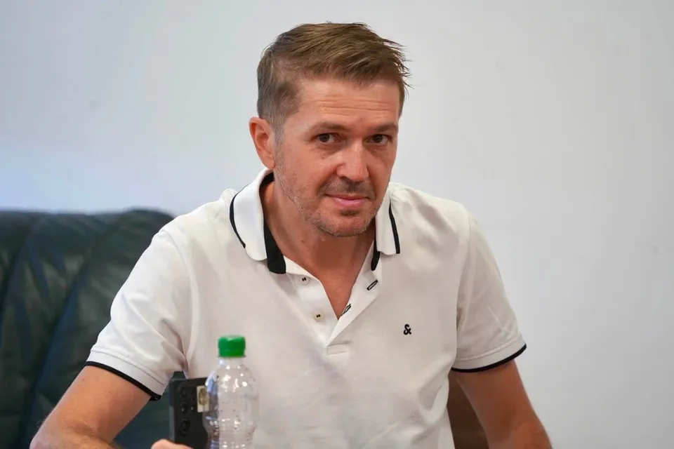 Un bărbat cu părul scurt și cămașă polo albă stă la o masă lângă o sticlă de apă transparentă.