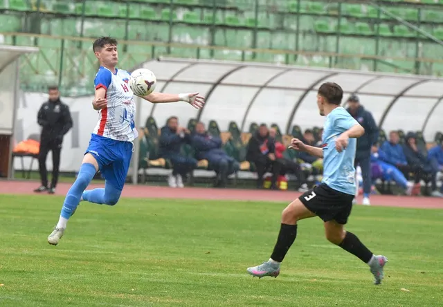 Un jucător în echipament albastru și roșu sare în aer pentru a lovi o minge de fotbal, în timp ce un alt jucător îl urmărește.