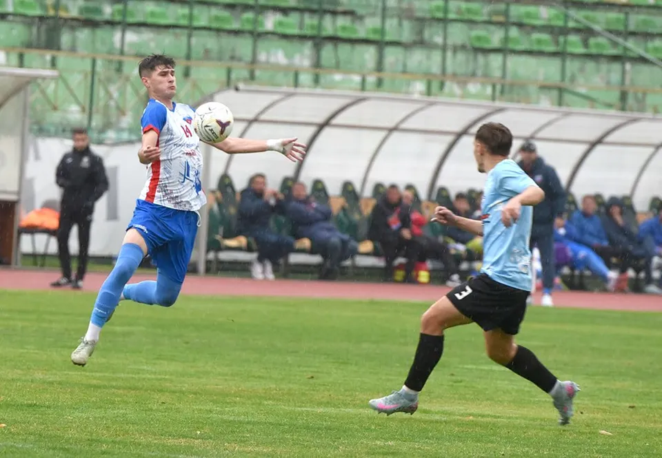 Un jucător în echipament albastru și roșu sare în aer pentru a lovi o minge de fotbal, în timp ce un alt jucător îl urmărește.
