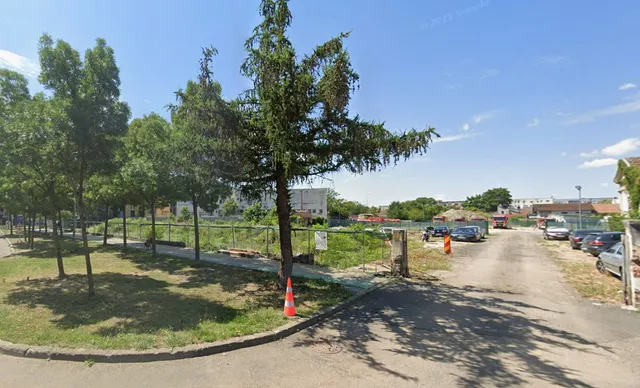 Un drum de asfalt trece pe lângă un teren cu iarbă și arbori, delimitat de un gard metalic sub un cer senin.