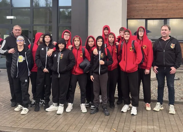 Un grup de adolescenți îmbrăcați în haine sportive negre și roșii stau în rând pe o suprafață pavată lângă o clădire.