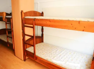 Două paturi de etaj din lemn cu saltele cu modele florale sunt amplasate lângă un dulap înalt într-o cameră cu pereți albi și podea de lemn.