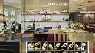 Rafturi de magazin conțin diverse modele de încălțăminte albe și colorate, expuse sub un afiș cu textul Golden Goose într-un spațiu cu suprafețe lucioase.