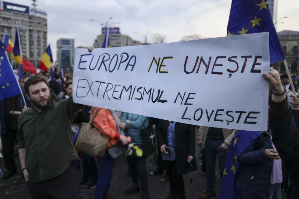 Un grup de persoane într-o mulțime strigă un protest, ținând un panou alb pe care scrie un text în limba română.