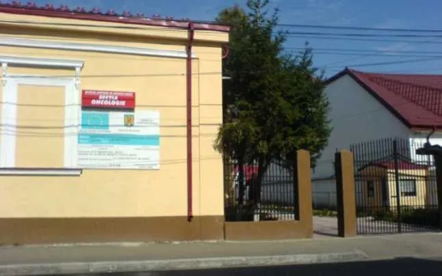 O clădire galbenă prezintă un panou informativ roșu și alb cu textul Secția Oncologie lângă un gard metalic și un copac.