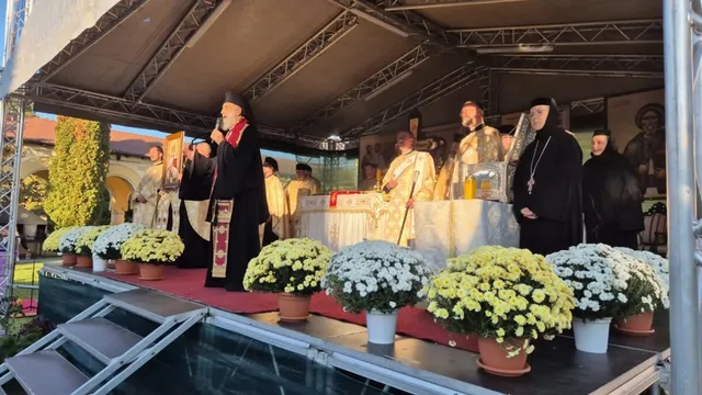 Câțiva bărbați în haine religioase negre și albe stau pe o platformă decorată cu flori albe și galbene sub un acoperiș metalic.