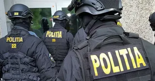 Trei polițiști în uniformă neagră cu inscripcții galbene sunt văzuți din spate, purtând căști și echipament tactic.