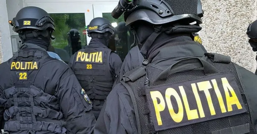 Trei polițiști în uniformă neagră cu inscripcții galbene sunt văzuți din spate, purtând căști și echipament tactic.