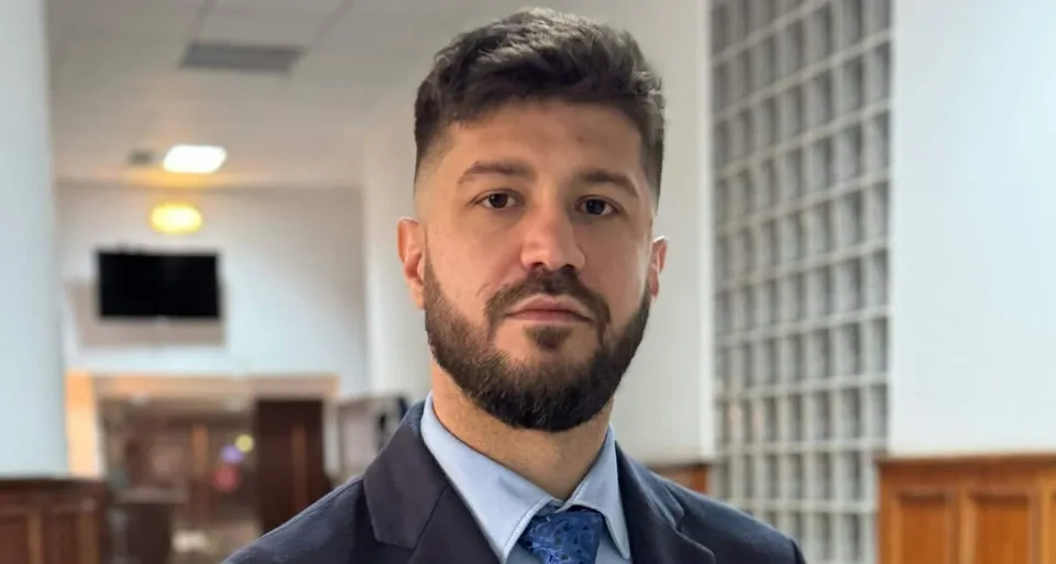 Un bărbat cu barbă și păr scurt, purtând sacou închis și cămașă albastră cu cravată, privește spre cameră într-un interior iluminat.