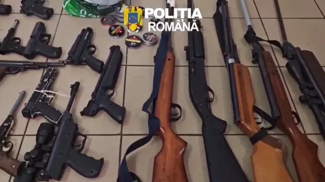 Diversele arme de foc negre și cu coci din lemn sunt așezate pe un podea de gresie alături de câteva obiecte rotunde mici.