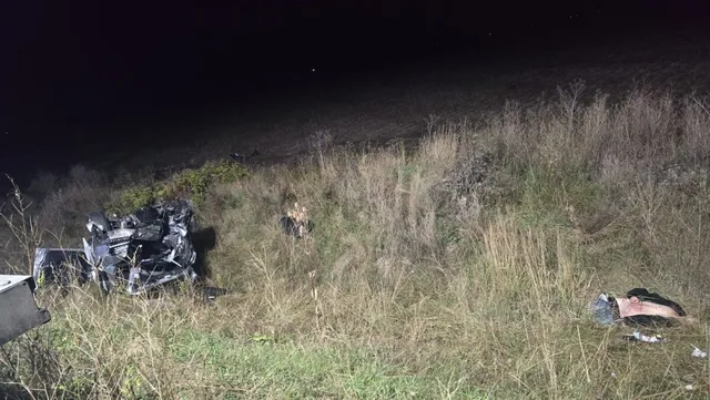 Un autoturism negru este răsturnat într-un teren cu iarbă înaltă sub lumină nocturnă.