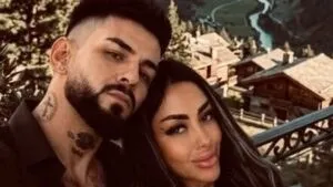 Un bărbat cu barbă și tatuaje pe gât stă lângă o femeie cu părul lung și machiaj accentuat în fața unui peisaj montan cu clădiri.