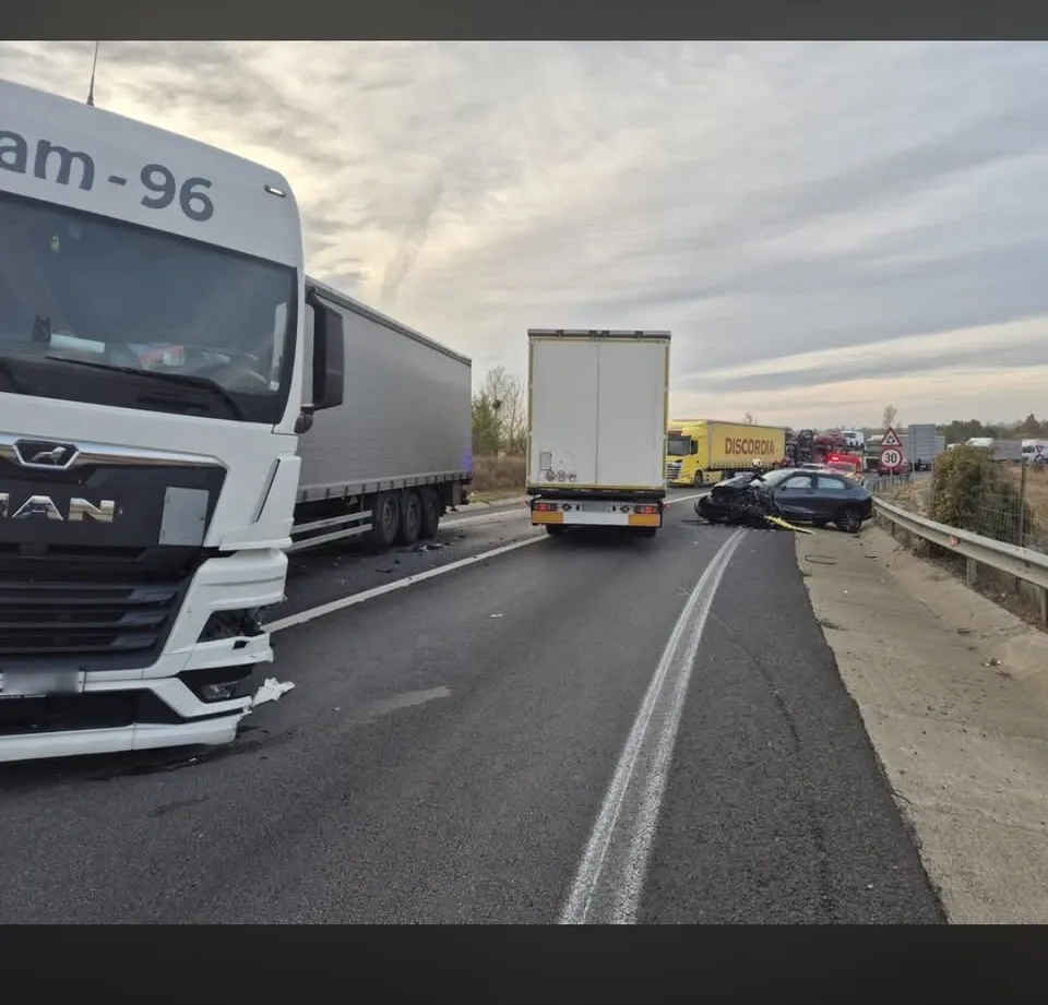 Un camion alb și un camion alb sunt oprite pe o autostradă lângă un autoturism negru de lângă o barieră metalică.