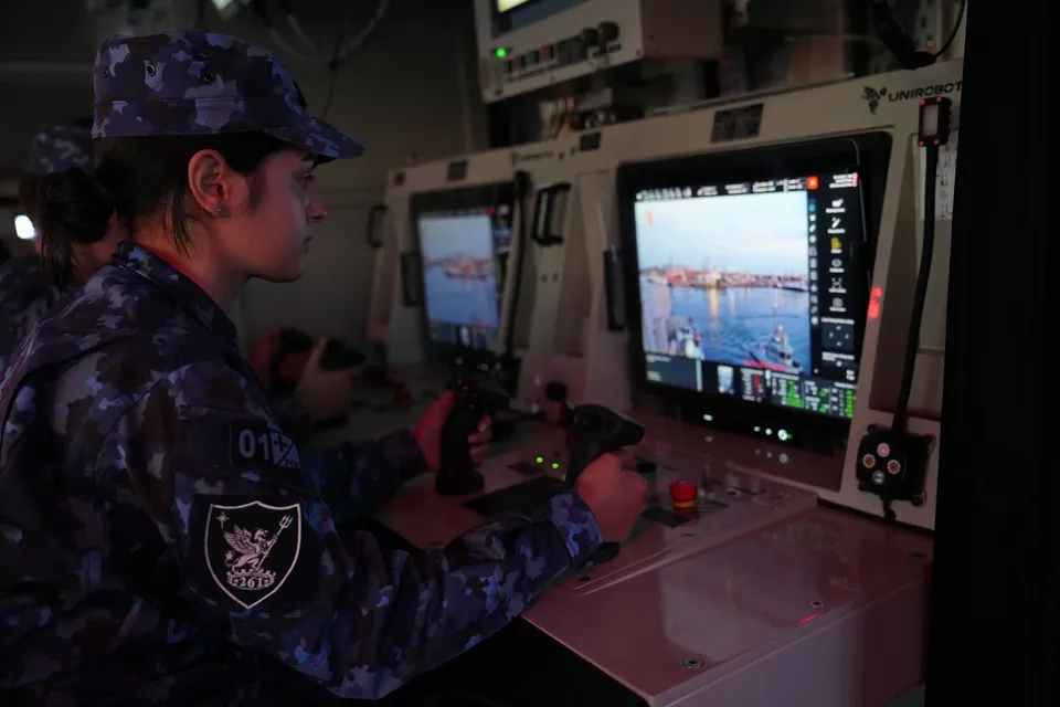 Un membru al personalului militar în uniformă camuflată albastru operează două dispozitive de control în fața unor monitoare digitale într-un interior tehnic.