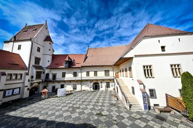 Un ansamblu de clădiri albe cu acoperișuri de țiglă roșie înconjoară o curte pavată cu pietre gri sub un cer albastru cu nori.