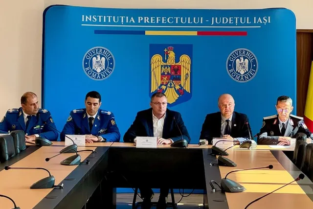 Cinci bărbați stau la o masă lungă în fața unui panou albastru pe care sunt imprimate embleme oficiale.