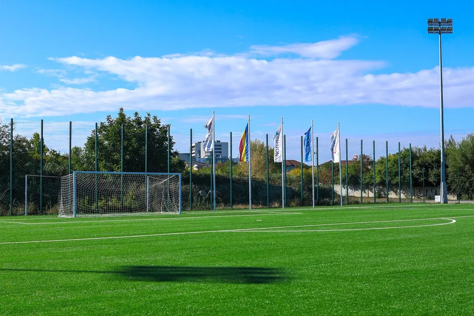 Un teren de fotbal cu iarbă artificială prezintă un por cu plasă și mai mulți stâlpi cu steaguri sub un cer albastru cu nori.