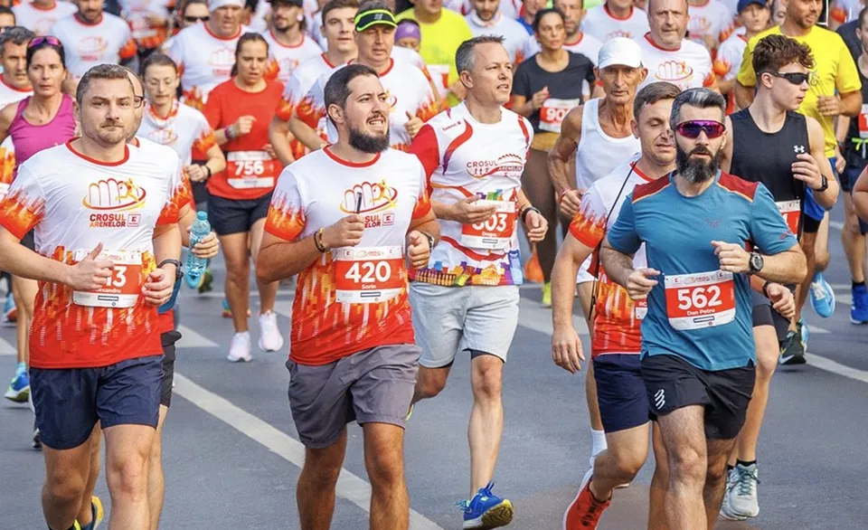Un grup de oameni în haine sportive participă la o cursă pe o pistă asfaltată, purtând numere de concurs pe tricouri.
