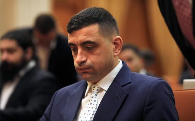 Un bărbat cu părul scurt și întunecat poartă un sacou albastru, o cămașă albă și o cravată albă cu modele geometrice mici.