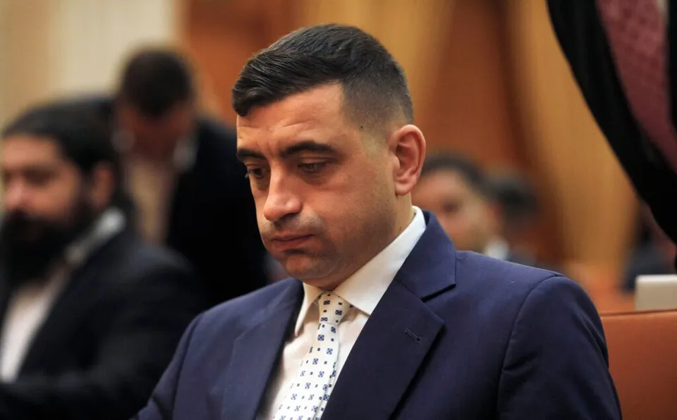 Un bărbat cu părul scurt și întunecat poartă un sacou albastru, o cămașă albă și o cravată albă cu modele geometrice mici.