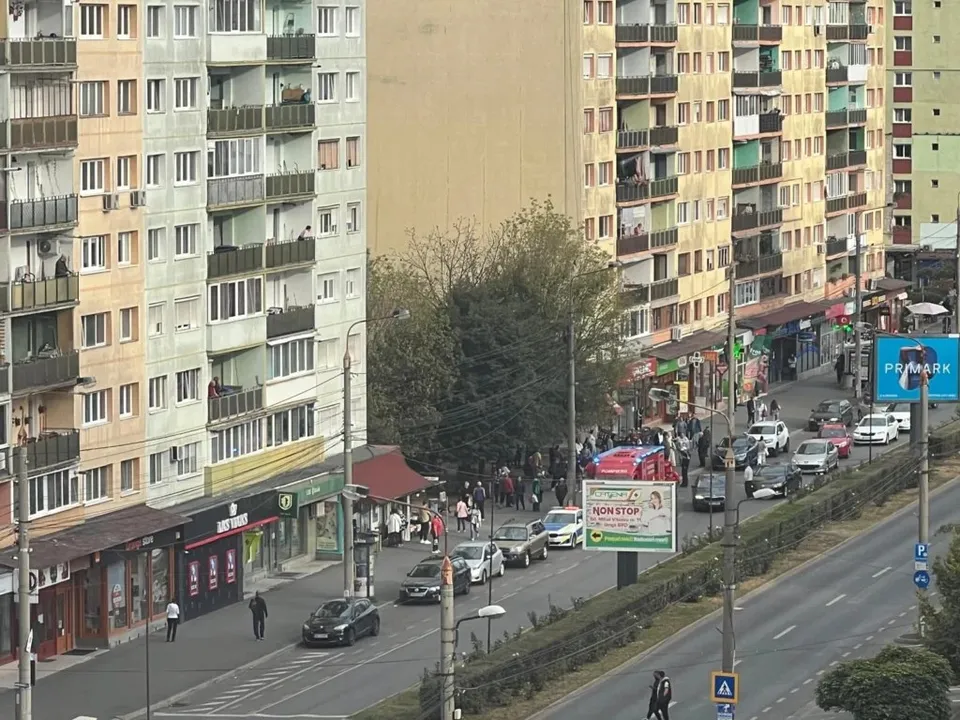 O stradă urbană cu clădiri înalte, mașini parcate și în mișcare, pietoni pe trotuar și un vehicul de urgență roșu.