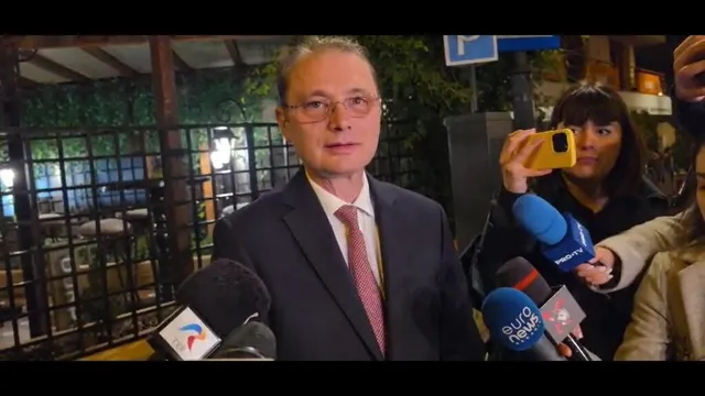 Un bărbat cu ochelari și cravată roz poartă un sacou închis la culoare în timp ce este înconjurat de microfoane și telefoane mobile.