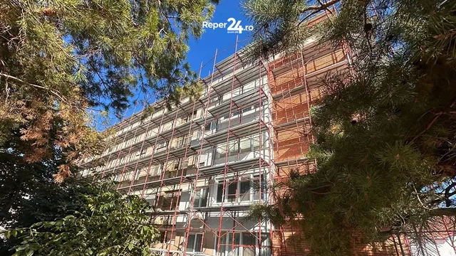O clădire înaltă este acoperită de schele metalice și este încadrată de ramuri de copaci verzi sub un cer albastru.