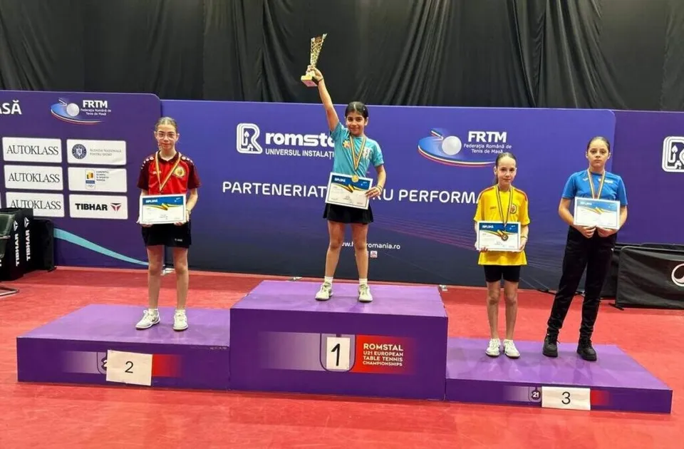 Trei fete stau pe podiumul de premiere, iar una ridică un trofeu deasupra capului în timp ce poartă o medalie și un certificat.