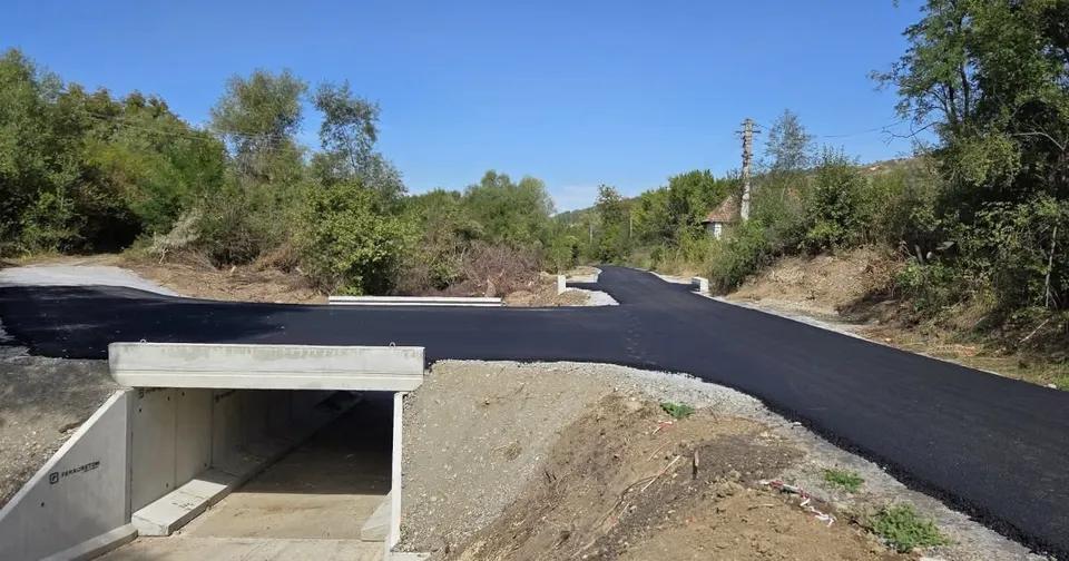 Un drum nou asfaltat cu un strat negru trece peste o structură de beton sub formă de tunel, înconjurat de vegetație și teren accidentat.
