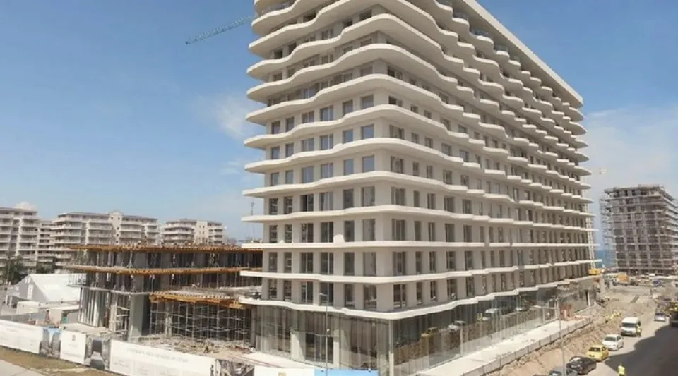 Un bloc de apartamente alb cu balcoane ondulate este situat lângă un șantier de construcții sub un cer senin.