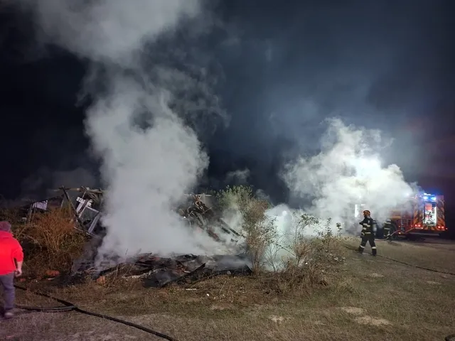 Fum alb se ridică deasupra unor resturi carbonizate într-un teren deschis, în prezența unui pompier și a unui vehicul de intervenție noaptea.