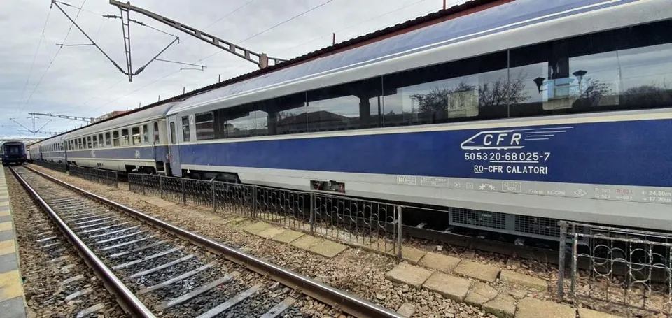 Un tren albastru și gri cu inscripția CFR stă pe o cale feroviară lângă un gard metalic sub un cer înnorat.