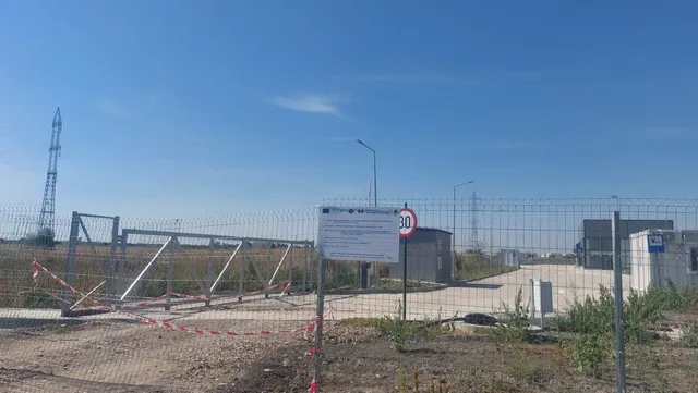 O clôtură metalică separă un teren cu pământ și vegetație scundă de o zonă pavată cu o clădire gri și un stâlp de iluminat.