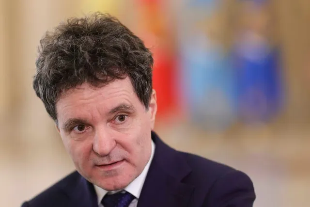 Un bărbat cu părul închis la culoare și scurt, purtând o jachetă închisă și o cămașă albă cu cravată, privește spre lateral.