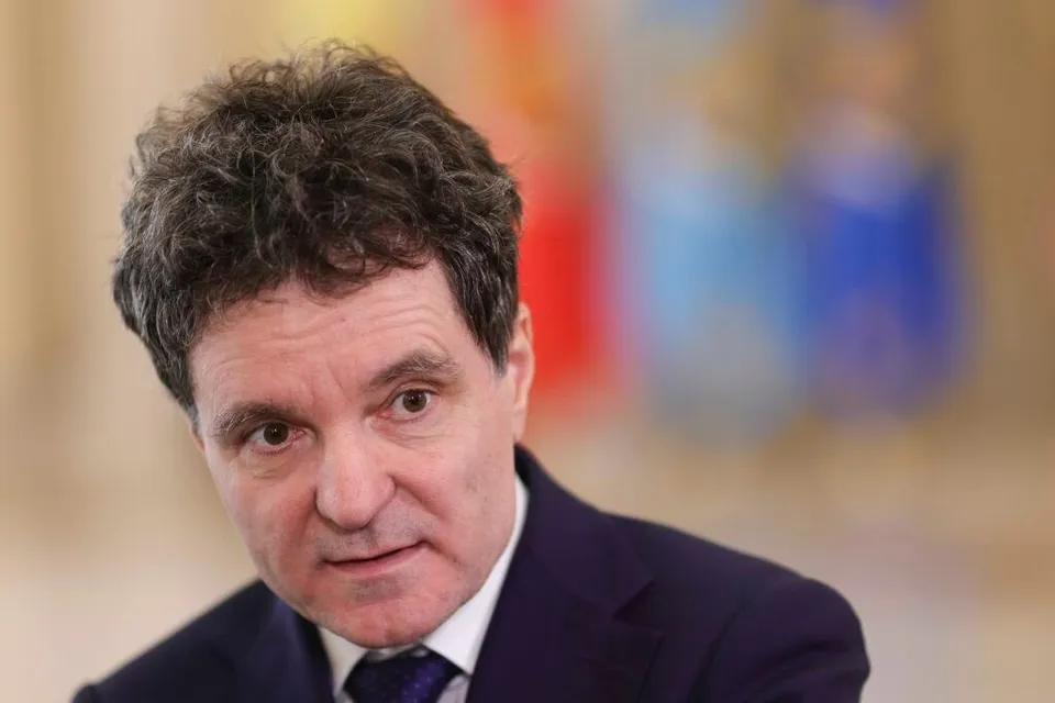 Un bărbat cu părul închis la culoare și scurt, purtând o jachetă închisă și o cămașă albă cu cravată, privește spre lateral.