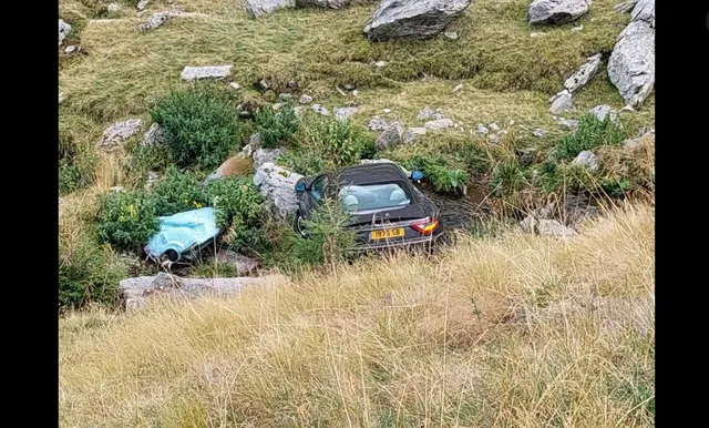 Un autoturism negru este situat într-o zonă cu iarbă și pietre, pe o pantă înclinată.