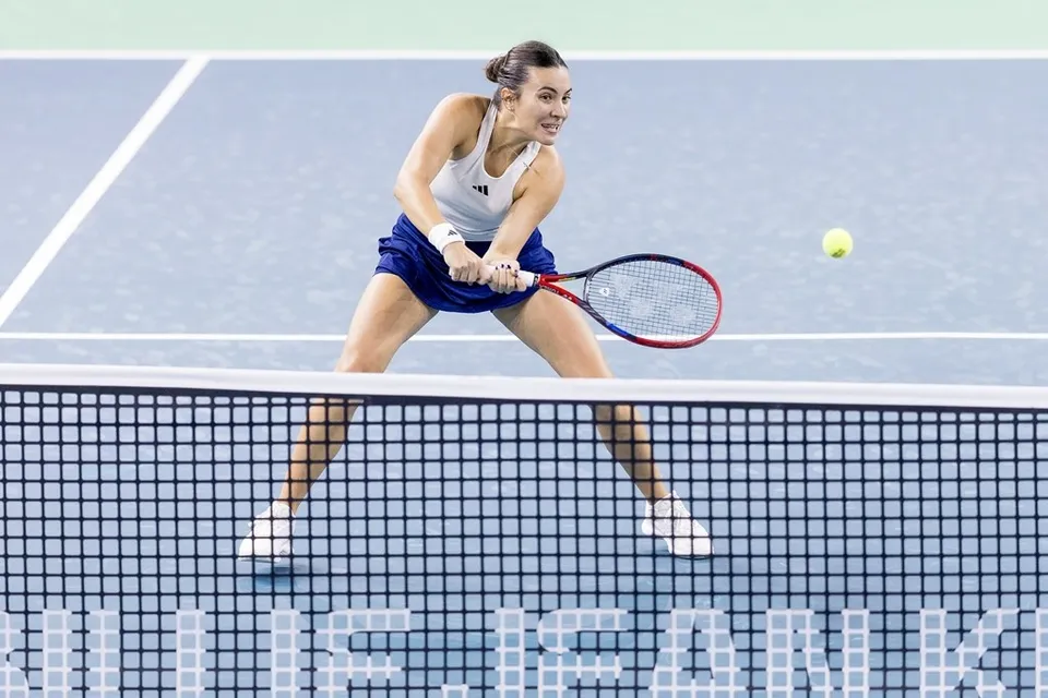 O femeie cu părul prins într-o coadă îmbrăcată într-un tricou alb și o fustă albastră lovește o minge galbenă cu o raketă pe un teren de tenis.