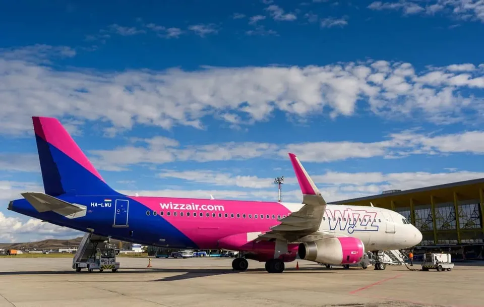 Un avion de pasageri roz și alb cu inscripția wizzair.com stă pe o pistă de aeroport sub un cer albastru cu nori albi.