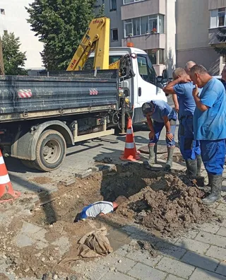 Un bărbat se află într-o groapă de pe drum, în timp ce alți bărbați stau lângă un camion cu braț hidraulic și un con portocaliu.