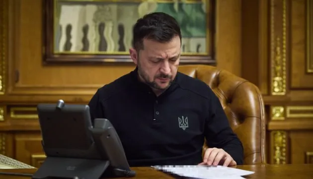 Un bărbat cu părul scurt și cămașă neagră cu un simbol pe piept stă la o masă de lemn și privește niște hârtii.