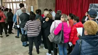 Un grup de persoane stă la coadă într-un spațiu interior cu podea de piatră și un fundal format din draperii roșii.