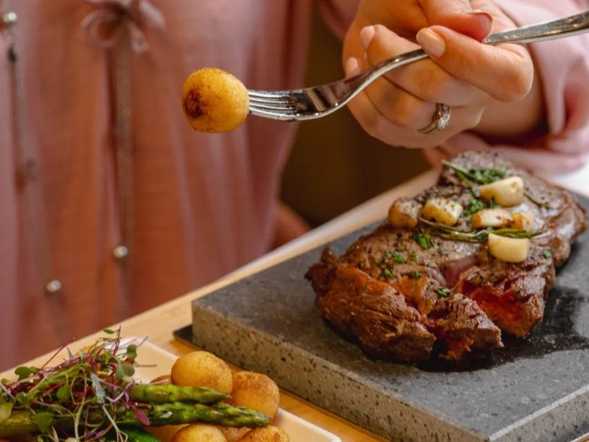 O persoană ține o furculiță cu un cartof mic, lângă o farfurie cu carne gătită pe o piatră și un sos de legume verzi.