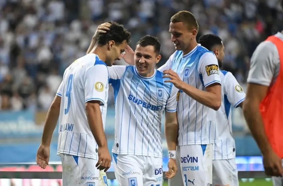 Cinci jucători de fotbal îmbrăcați în echipamente albe cu dungi albastre stau grupați pe un teren de sport, în timp ce un spectator este parțial vizibil.