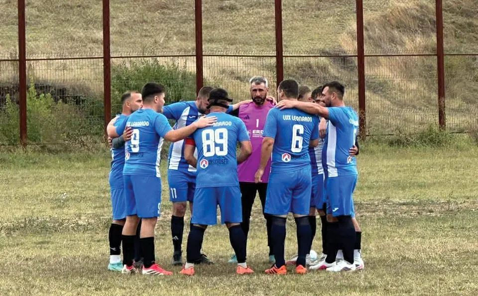 Un grup de fotbaliști în echipamente albastre stau într-un cerc pe un teren de fotbal, cu brațele pe umerii colegilor.
