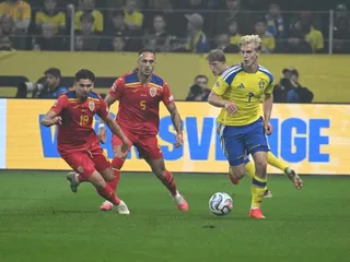 Trei jucători de fotbal sunt pe teren de iarbă, unul purtând echipament galben și albastru, iar ceilalți doi purtând echipament roșu și alb.