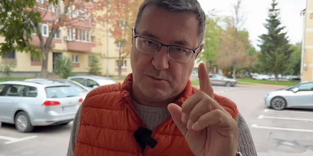 Un bărbat cu ochelari și vestă portocalie vorbește și arată cu degetul într-un cadru exterior cu mașini și clădiri.