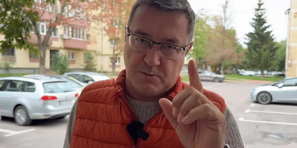Un bărbat cu ochelari și vestă portocalie vorbește și arată cu degetul într-un cadru exterior cu mașini și clădiri.